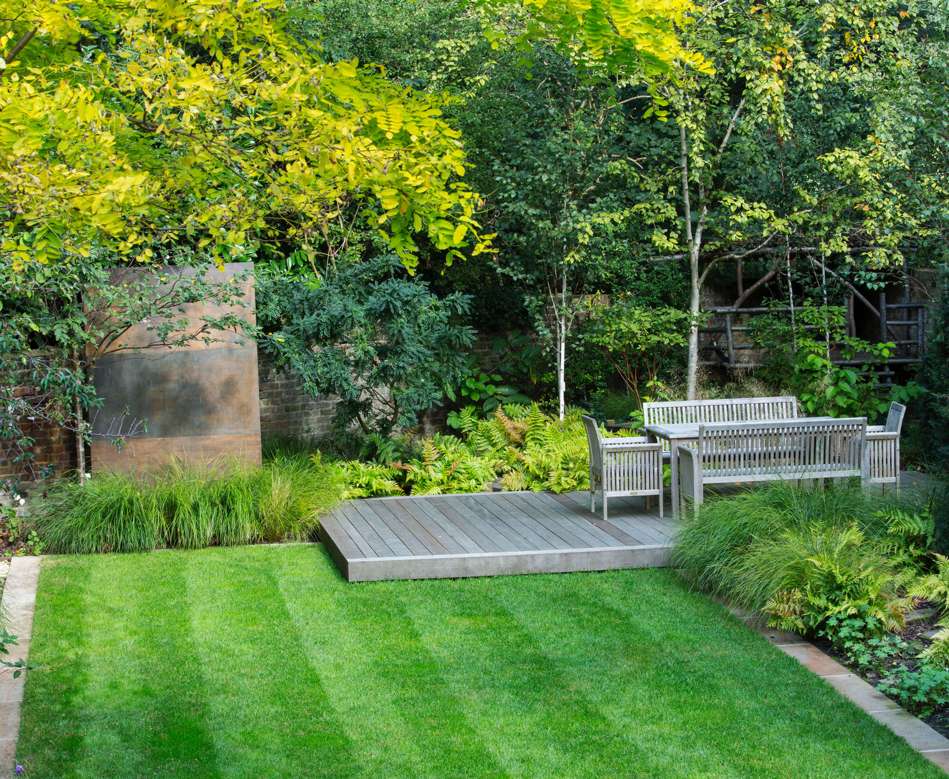 The raised hardwood timber deck floats above the lawn, just in front of the hidden play house, overlooking Hampstead Heath. The woodland planting moves towards the lawn, where a feature bronze panel stands. Some grasses soften the edges of the turf, making it disappear.