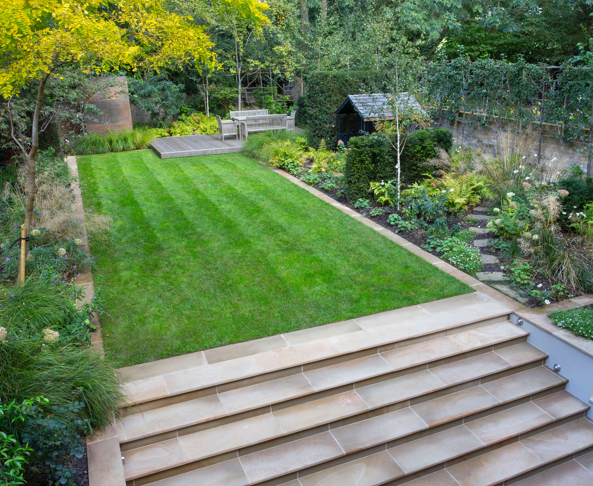 The rectangular lawn is accessed from the lower patio using long, wide yorkstone steps. Some mixed borders on either side make the boundaries disappear, with the addition of some pleached trees. At the back, the woodland planting merges with the existing forest, whilst an old Gleditsia with yellow leaves becomes a main focal point to enjoy from the house.