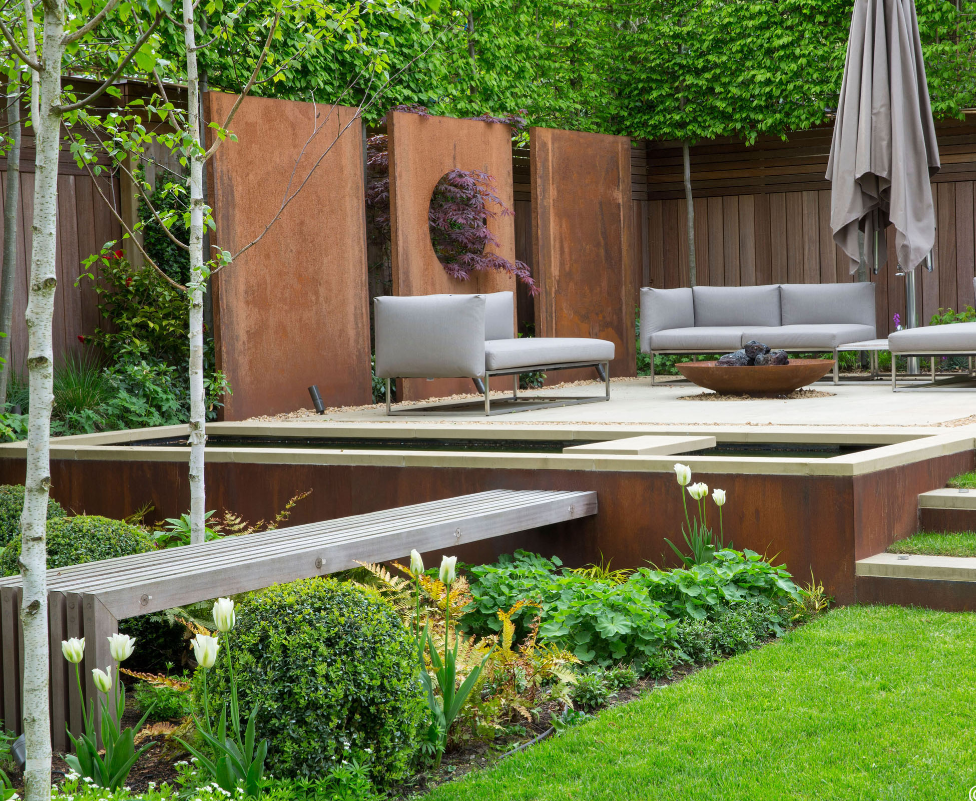 A view of the Highgate garden showing the comfortable seating area, borded by a bespoke water feature and some corten steel panels. Some tall pleached trees provide intimacy from the neighbours.