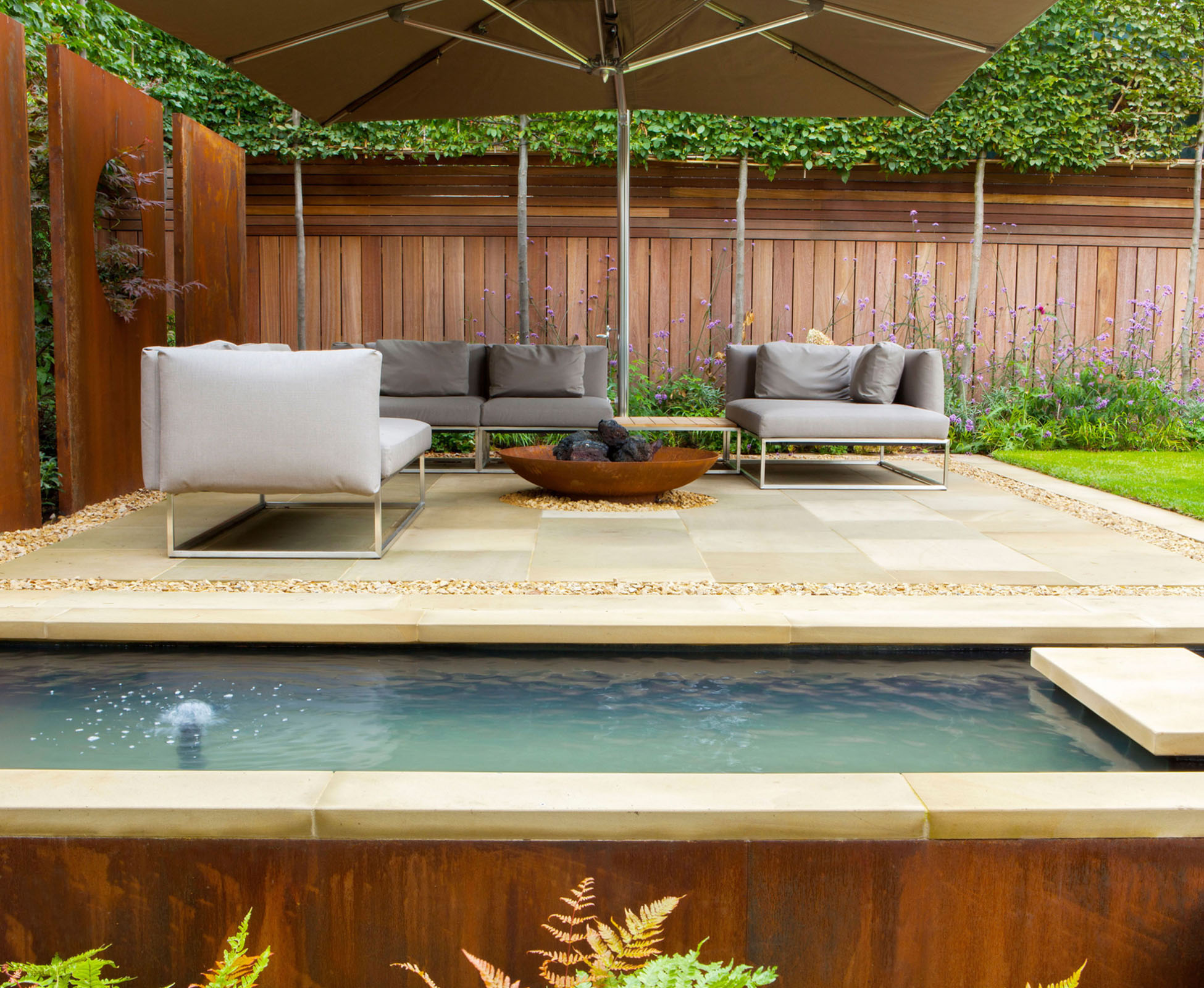 Some contemporary furniture compliment the corten features and fire bowl. The warm colours of the beige sandstone and rusted steel contrast with the green pleached trees and with the clear blue water feature in this Highgate garden.