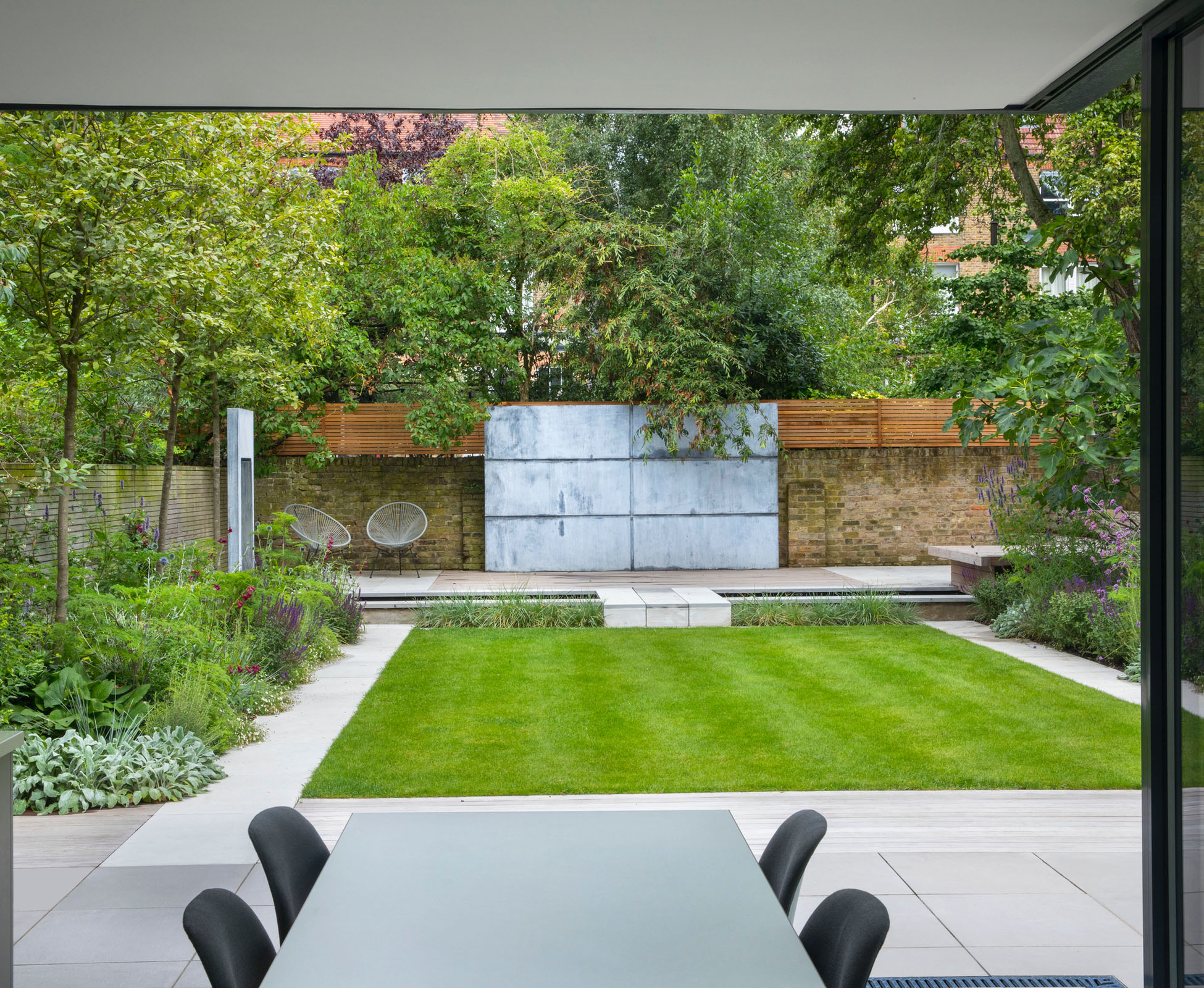 This Islington garden with its rectangular lawn is designed with a strong focal view out of the house to a zinc panel and water feature. A sandstone bridge let us cross the gap to a decked seating area.